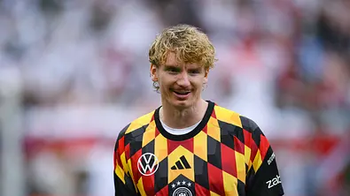 Ein Fußballspieler der deutschen Nationalmannschaft steht auf dem Spielfeld und trägt ein auffällig gemustertes Trikot in Schwarz, Rot und Gelb. Er blickt lächelnd zur Seite, während im Hintergrund die Zuschauertribünen unscharf zu erkennen sind. Die Aufnahme entstand in einem Stadion vor oder nach einem Spiel. - Foto: IMAGO / Ulmer/Teamfoto Ein Fußballspieler der deutschen Nationalmannschaft steht auf dem Spielfeld und trägt ein auffällig gemustertes Trikot in Schwarz, Rot und Gelb. Er blickt lächelnd zur Seite, während im Hintergrund die Zuschauertribünen unscharf zu erkennen sind. Die Aufnahme entstand in einem Stadion vor oder nach einem Spiel. - Foto: IMAGO / Ulmer/Teamfoto