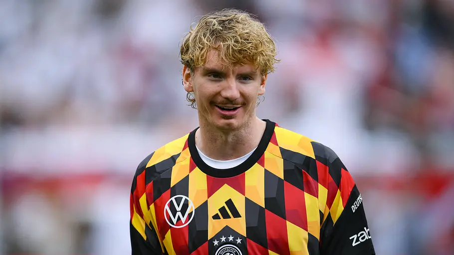 Nick Woltemade Ein Fußballspieler der deutschen Nationalmannschaft steht auf dem Spielfeld und trägt ein auffällig gemustertes Trikot in Schwarz, Rot und Gelb. Er blickt lächelnd zur Seite, während im Hintergrund die Zuschauertribünen unscharf zu erkennen sind. Die Aufnahme entstand in einem Stadion vor oder nach einem Spiel.