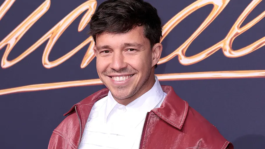 Nico Santos lächelt in roter Lederjacke und weißem Hemd vor einem dunklen Hintergrund mit goldfarbenem Schriftzug.