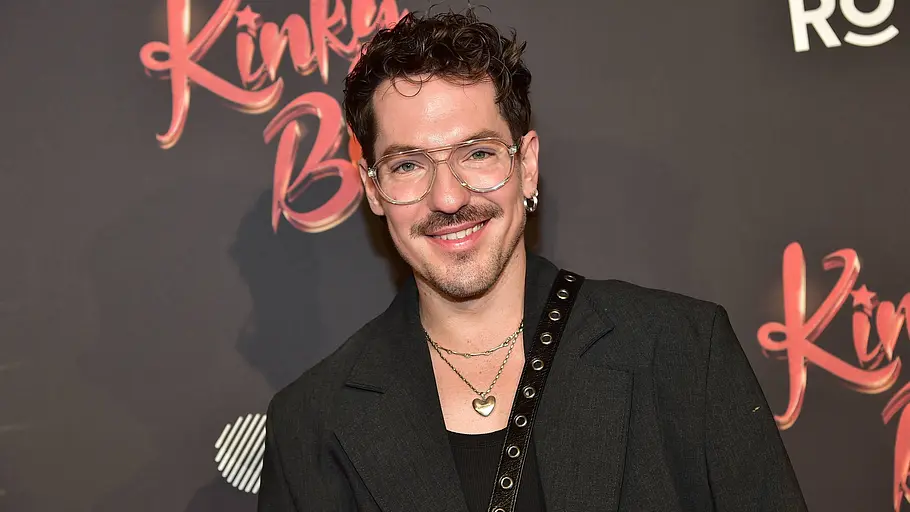 Nico Stank lächelt mit Brille und Schnurrbart vor einer Logowand. Er trägt einen dunklen Blazer und eine Kette mit Herzanhänger.