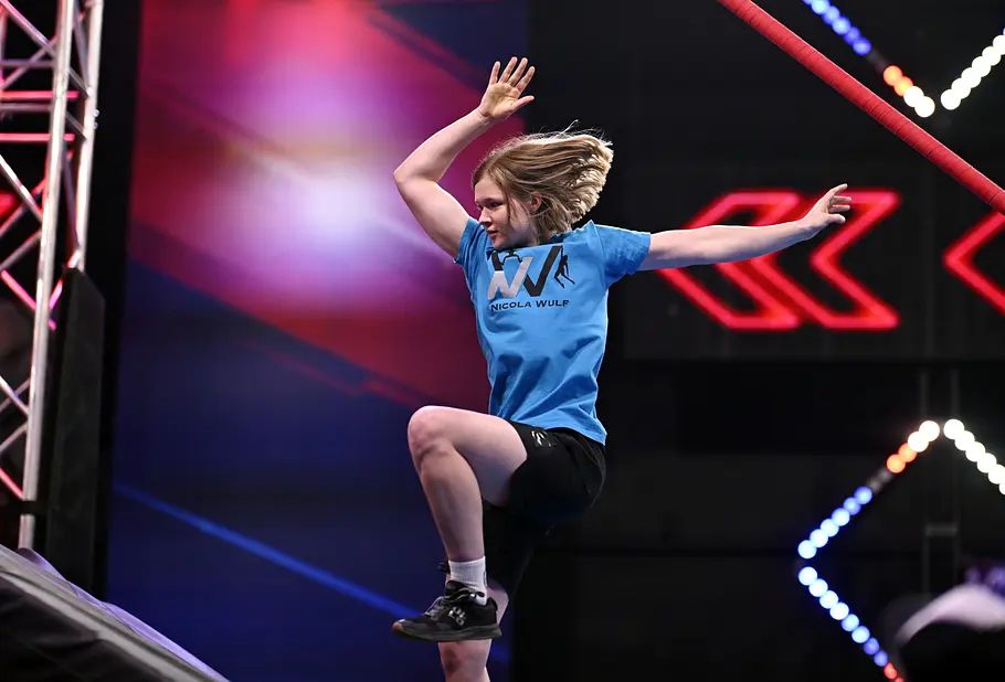 Nicola Wulf ist Teilnehmer bei Ninja Warrior Germany 2026 Nicola Wulf hebt im Hindernisparcours ein Knie und breitet die Arme aus. Er trägt ein blaues T-Shirt und schwarze Shorts.
