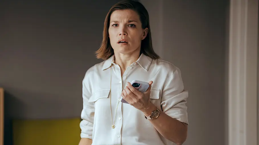 Nicole ist die Ehefrau des Immobilienmanagers Benedikt Renner. Nicole Renner (Mona Kospach) hält in einer weißen Bluse ein Smartphone vor der Brust und blickt mit leicht geöffnetem Mund.