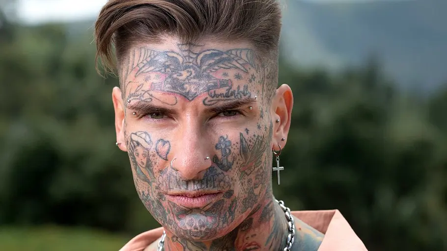 Nikola Glumac alias Nikola Grey Nikola Glumac alias Nikola Grey blickt mit tätowiertem Gesicht, Piercings und einem Kreuzohrring in die Kamera.