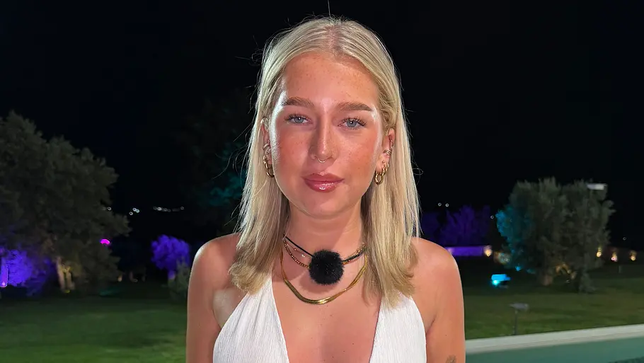 Nina, 21 Eine blonde Frau in weißem Oberteil und kurzem Rock steht nachts vor einem Pool mit Schwimmringen und beleuchteten Bäumen.