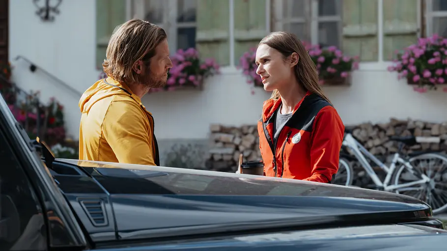 Markus in gelber und Nina in roter Jacke stehen sich hinter einem dunklen Geländewagen vor einem Haus mit Blumenkästen gegenüber.