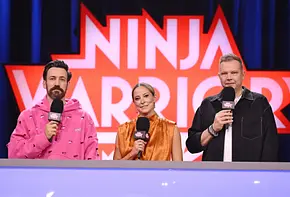 Jan Köppen, Anna Fleischhauer und Wolff Fuss stehen mit Mikrofonen vor dem „Ninja Warrior“-Logo hinter einem Pult. - Foto: Foto: RTL / Markus Hertrich Jan Köppen, Anna Fleischhauer und Wolff Fuss stehen mit Mikrofonen vor dem „Ninja Warrior“-Logo hinter einem Pult. - Foto: Foto: RTL / Markus Hertrich