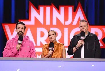 Jan Köppen, Anna Fleischhauer und Wolff Fuss stehen mit Mikrofonen vor dem „Ninja Warrior“-Logo hinter einem Pult. - Foto: Foto: RTL / Markus Hertrich