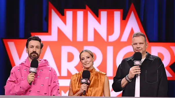 Jan Köppen, Anna Fleischhauer und Wolff Fuss stehen mit Mikrofonen vor dem „Ninja Warrior“-Logo hinter einem Pult. - Foto: Foto: RTL / Markus Hertrich Jan Köppen, Anna Fleischhauer und Wolff Fuss stehen mit Mikrofonen vor dem „Ninja Warrior“-Logo hinter einem Pult. - Foto: Foto: RTL / Markus Hertrich