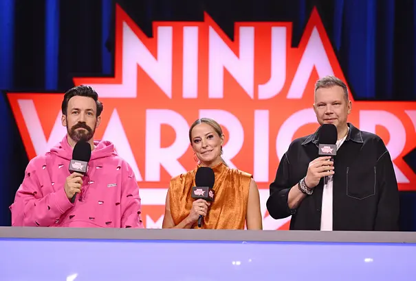 Jan Köppen, Anna Fleischhauer und Wolff Fuss stehen mit Mikrofonen vor dem „Ninja Warrior“-Logo hinter einem Pult. - Foto: Foto: RTL / Markus Hertrich Jan Köppen, Anna Fleischhauer und Wolff Fuss stehen mit Mikrofonen vor dem „Ninja Warrior“-Logo hinter einem Pult. - Foto: Foto: RTL / Markus Hertrich