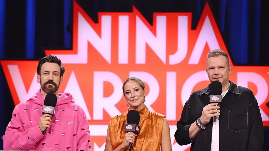 „Ninja Warrior“-Teilnehmer 2026 Jan Köppen, Anna Fleischhauer und Wolff Fuss stehen mit Mikrofonen vor dem „Ninja Warrior“-Logo hinter einem Pult.