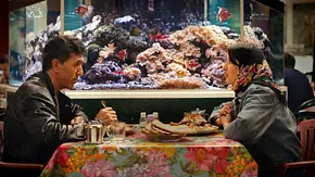 Ein Mann und eine Frau sitzen in einem Restaurant am Tisch. Im Hintergrund ist ein Aquarium zu sehen. - Foto: Virginie Surdej