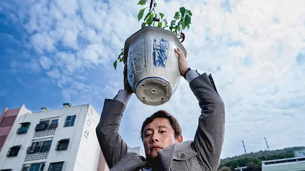 Der Hauptdarsteller aus „No Other Choice“ steht an einer Dachkante und hält einen Blumentopf über seinem Kopf. - Foto: IMAGO / Landmark Media Der Hauptdarsteller aus „No Other Choice“ steht an einer Dachkante und hält einen Blumentopf über seinem Kopf. - Foto: IMAGO / Landmark Media