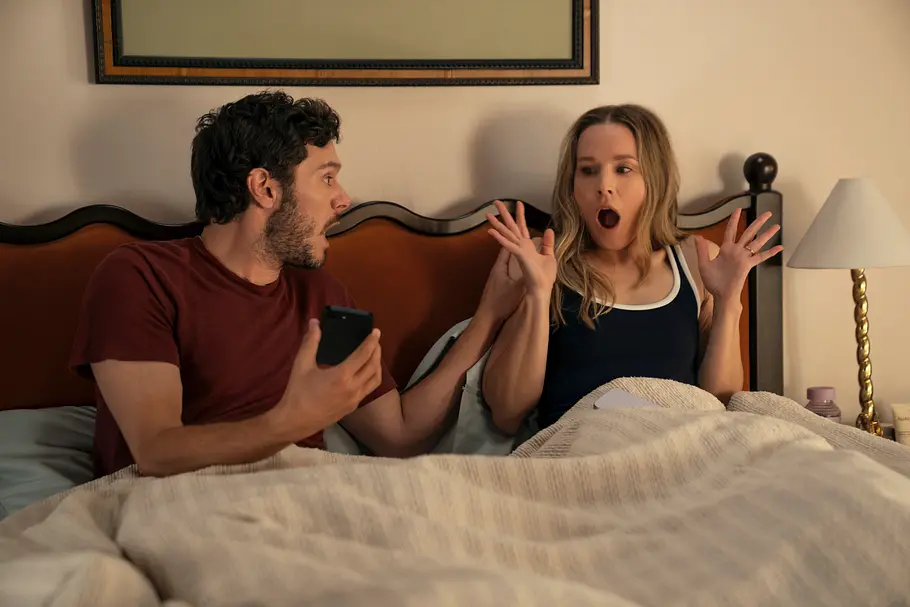 Joanne und Noah heiraten am Strand Noah und Joanne sitzen im Bett. Er hält ein Smartphone, sie hebt mit geöffnetem Mund und gespreizten Fingern die Hände.