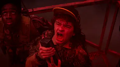 Dustin hält mit offenem Mund ein Funkgerät. Neben ihm steht Lucas im roten Licht vor einem Metallgeländer. - Foto: Netflix Dustin hält mit offenem Mund ein Funkgerät. Neben ihm steht Lucas im roten Licht vor einem Metallgeländer. - Foto: Netflix
