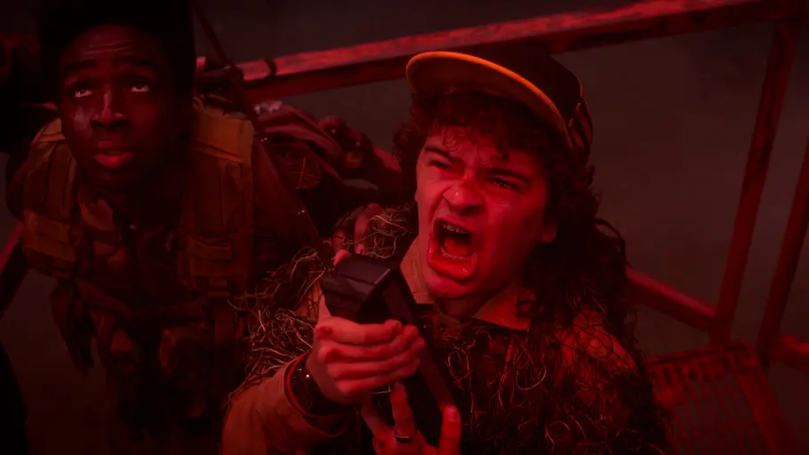 Noch magischer als die Welt von „Stranger Things“ ist das Denken der Fans. Dustin hält mit offenem Mund ein Funkgerät. Neben ihm steht Lucas im roten Licht vor einem Metallgeländer.