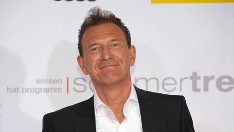 Moderator Norbert Lehmann nett lächelnd vor einer Fotowand. Er trägt ein schwarzes Jackett, sowie ein weißes Hemd. - Foto:  IMAGO / POP-EYE