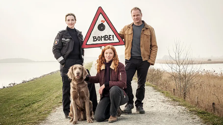 Nord bei Nordwest: Blindgänger Das Team aus „Nord bei Nordwest“ steht mit einem Hund und einem Bombenwarnungsschild auf einem Deich.