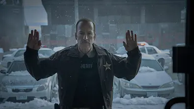 Bob Odenkirk steht im Film „Normal“ als Sheriff mit erhobenen Wänden vor einem verschneiten Parkplatz.  - Foto: Leonine