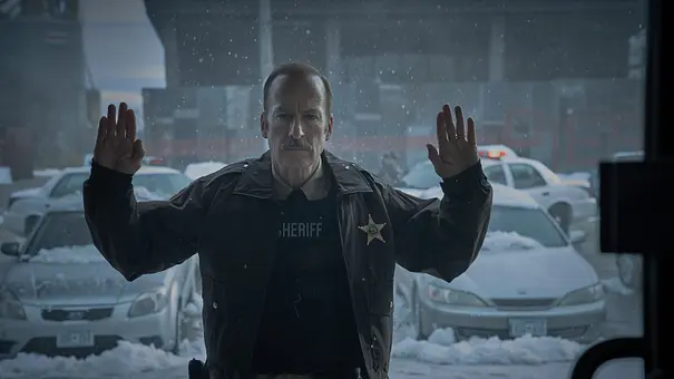 Bob Odenkirk steht im Film „Normal“ als Sheriff mit erhobenen Wänden vor einem verschneiten Parkplatz.  - Foto: Leonine