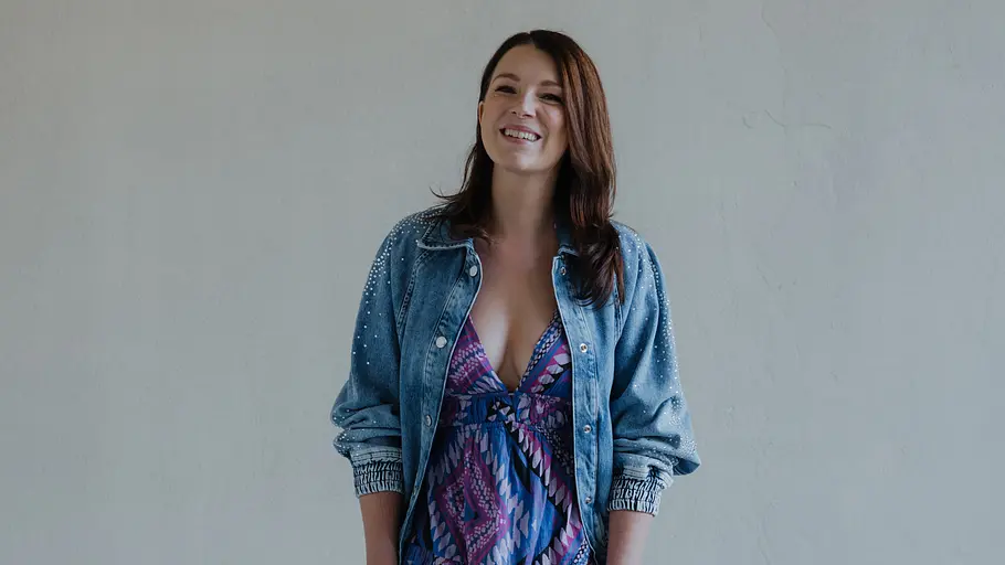 Nun rätseln Fans, wie es mit Lilly Seefeld bei GZSZ weitergeht. Iris Mareike Steen steht lächelnd in Jeansjacke, bunt gemustertem Kleid und fliederfarbenen Pumps vor einer hellgrauen Wand.