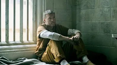 Sam Worthington sitzt in Gefängnisklamotten in einer Gefängniszelle. - Foto: Christos Kalohoridis/NETFLIX © 2025 Sam Worthington sitzt in Gefängnisklamotten in einer Gefängniszelle. - Foto: Christos Kalohoridis/NETFLIX © 2025