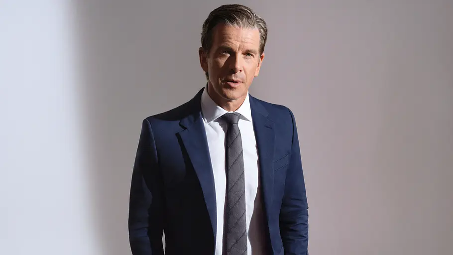 Ob sich die Kaulitz-Brüder besser schlagen als Markus Lanz? Markus Lanz steht im dunkelblauen Anzug mit weißem Hemd und grauer Krawatte vor einer hellen Wand, die Hände in den Taschen.