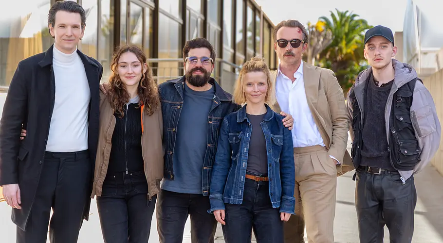 Gruppenfoto von sieben erwachsenen Personen, die nebeneinander im Freien stehen. Sie tragen überwiegend legere Kleidung wie Jacken, Jeans und Blazer. Im Hintergrund sind ein modernes Gebäude mit Glasfronten sowie Palmen zu sehen, bei sonnigem Tageslicht.