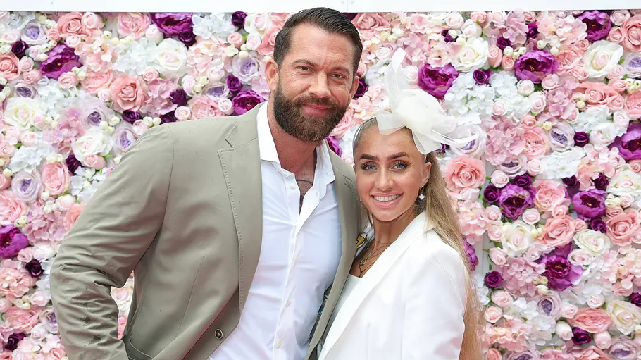 Oliver Sanne und Jil Rock Oliver Sanne im hellen Anzug und Jil Rock in Weiß mit großem Haarschmuck posieren lächelnd vor einer bunten Blumenwand.