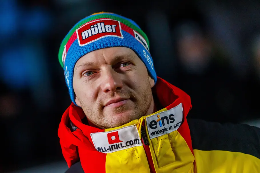 Deutscher Wintersportler mit Mütze und Jacke in Nationalfarben blickt konzentriert in die Kamera, Sponsorenlogos auf Kleidung sichtbar.