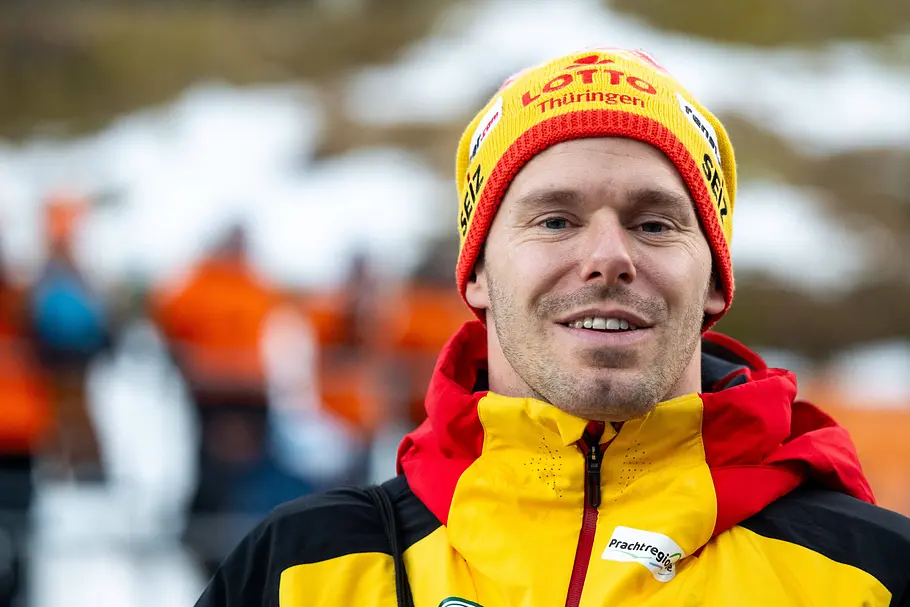 Porträt eines Mannes in gelb-roter Winterjacke und passender Mütze, der draußen in winterlicher Umgebung steht. Im Hintergrund sind unscharf weitere Personen und Schnee zu sehen.