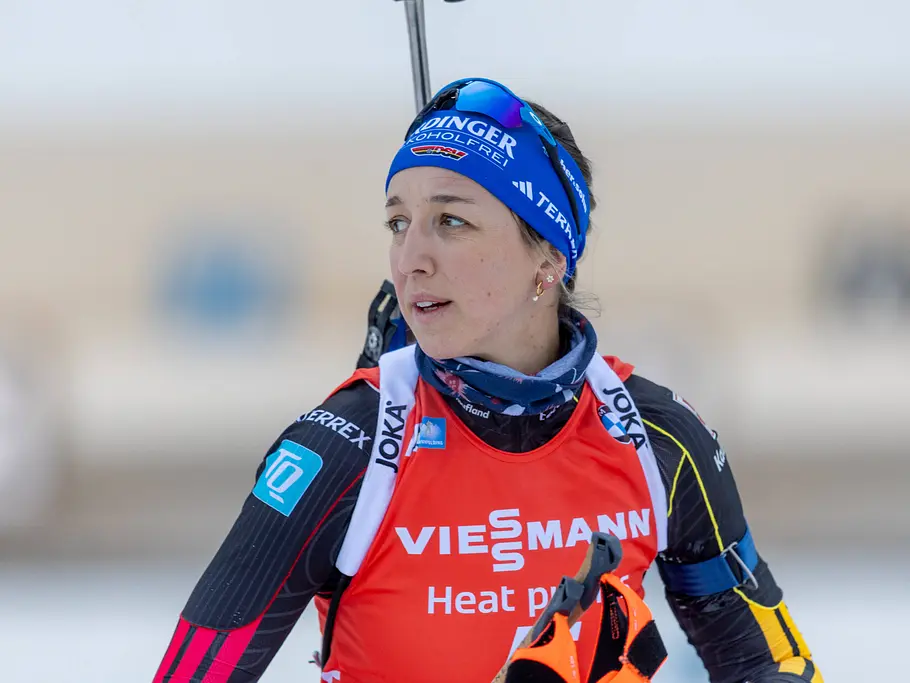 Sportlerin in rotem Wettkampfanzug mit Stirnband und Skibrille auf dem Kopf, die bei winterlichen Bedingungen im Freien steht und zur Seite blickt. Im Hintergrund ist eine verschneite, unscharfe Umgebung zu sehen.