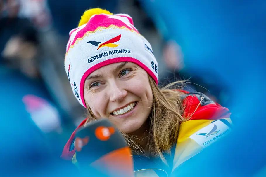 Bobpilotin Laura Nolte lächelt in die Kamera, trägt eine Mütze und Jacke in deutschen Nationalfarben mit Sponsorenlogos.