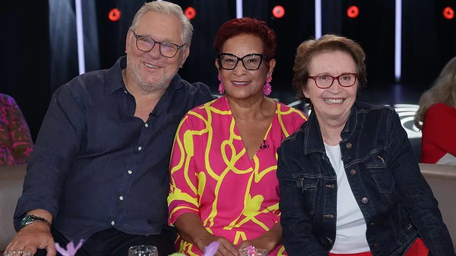 OmaLiebe e.V. Drei Personen von OmaLiebe e.V. sitzen eng nebeneinander auf einem Sofa im Studio einer Talkshow und lächeln in die Kamera. Links ein Mann mit grauem Haar und Brille in einem dunklen Hemd, in der Mitte eine Frau mit kurzen roten Haaren, auffälliger Brille und einem pink‑gelb gemusterten Kleid, rechts eine Frau mit kurzer brauner Frisur, roter Brille, Jeansjacke und roter Hose. Auf dem Tisch davor stehen Wassergläser und bunte Blumen, im Hintergrund sind weitere Gäste und Studiolichter zu sehen.