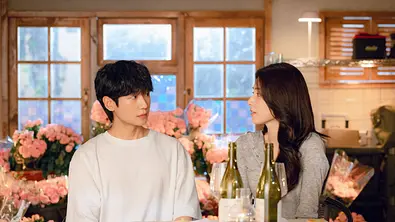 Tae-ha und Eun-sae sitzen an einem Tisch mit Wein. Hinter ihnen sich viele rosa Blumen. - Foto: Courtesy of Netflix