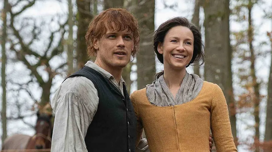 Jamie und Claire in einem Wald. Sie umarmen sich von der Seite und lächeln beide.
