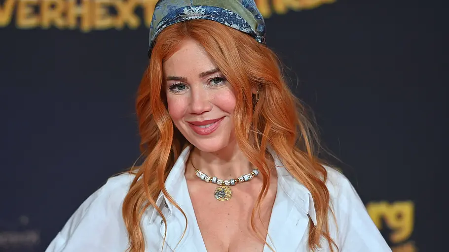 Palina Rojinski lächelt mit offenem rotem Haar, gemustertem Kopftuch, weißer Bluse und Halskette vor dunklem Hintergrund.