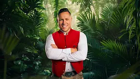 Patrick Romer steht mit roter Weste und weißem Hemd, sowie verschränkten Armen, lächelnd vor einer Dschungeloptik. - Foto: RTL / Hintergrund KI-generiert Patrick Romer steht mit roter Weste und weißem Hemd, sowie verschränkten Armen, lächelnd vor einer Dschungeloptik. - Foto: RTL / Hintergrund KI-generiert