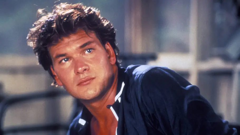 Patrick Swayze in „Dirty Dancing“. Patrick Swayze mit dunklen, zerzausten Haaren und offenem dunkelblauem Hemd blickt schräg nach oben. Der Hintergrund ist unscharf.
