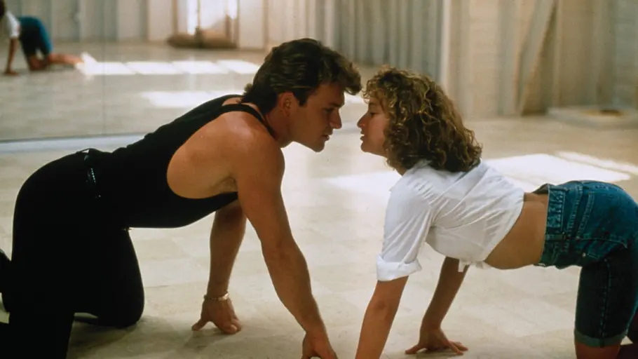 Patrick Swayze und Jennifer Grey. Patrick Swayze in Schwarz und Jennifer Grey in weißer Bluse und Jeansshorts stehen auf allen vieren, die Gesichter nah beieinander.