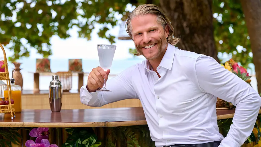 Paul Janke Paul Janke sitzt lächelnd im weißen Hemd an einer Bar im Freien und hält ein Stielglas. Auf der Theke stehen Früchte und Getränke.