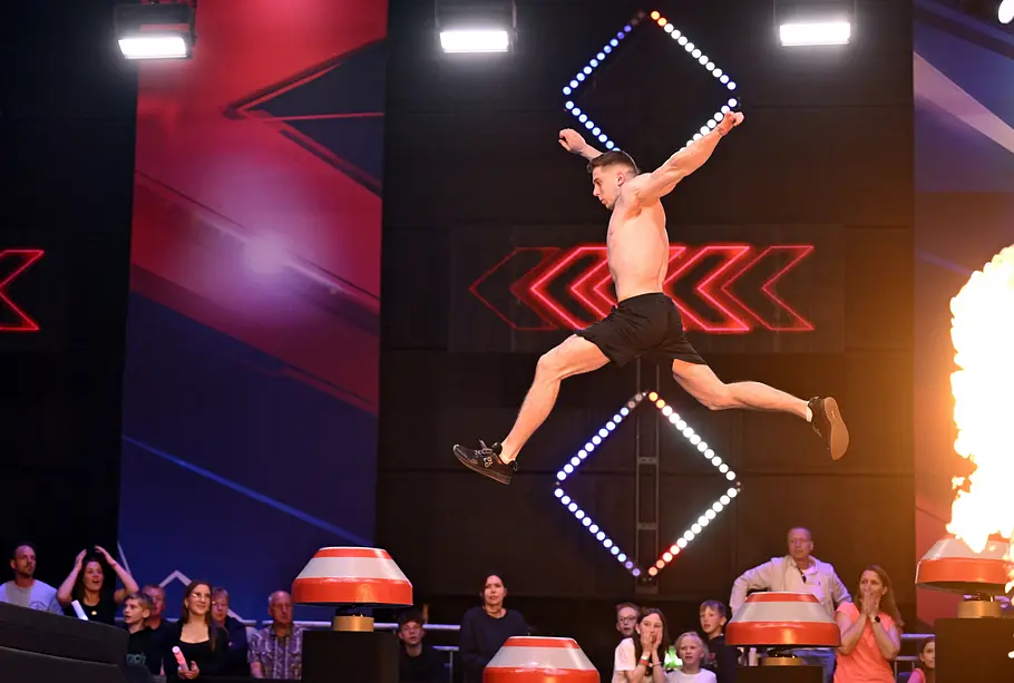 Paul Stöckel ist Teilnehmer bei Ninja Warrior Germany 2026 Paul Stöckel springt mit freiem Oberkörper und ausgebreiteten Armen über rote Hindernisse. Im Hintergrund schaut Publikum zu.