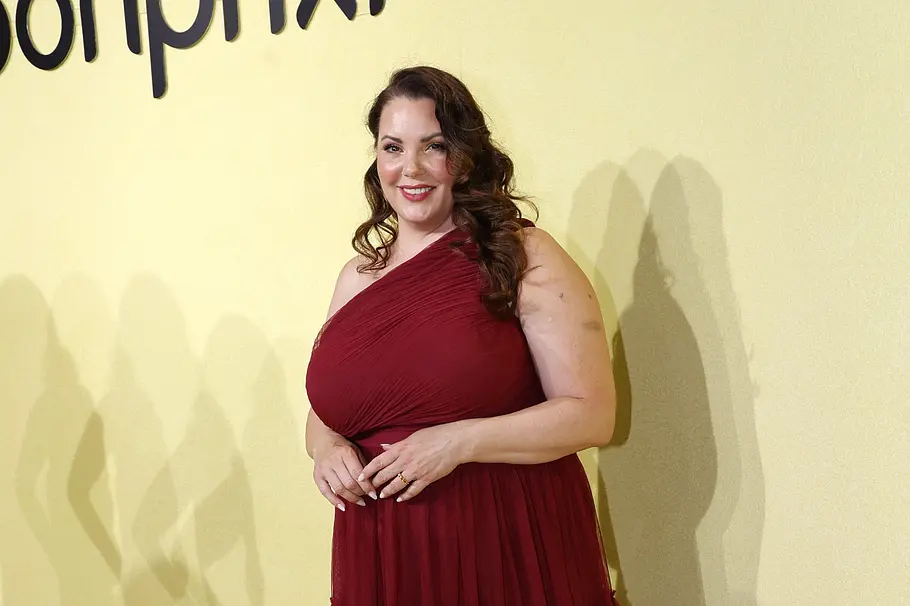 Paula Lambert mit langen braunen Locken steht in einem dunkelroten, einseitig schulterfreien Kleid vor gelber Wand.