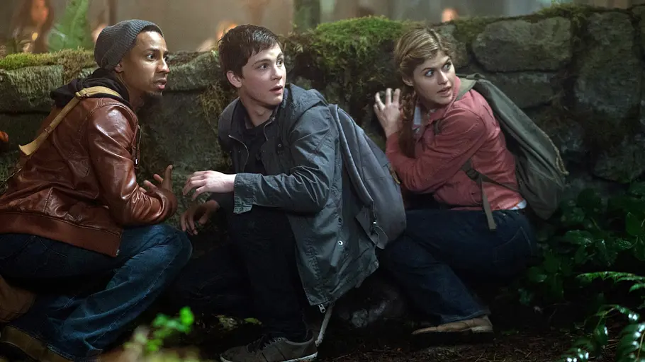Percy Jackson, Annabeth Chase und Grover Underwood ducken sich in einer Waldszene hinter einer Steinmauer und blicken alarmiert in verschiedene Richtungen – Szene aus dem Film „Percy Jackson – Im Bann des Zyklopen“.
