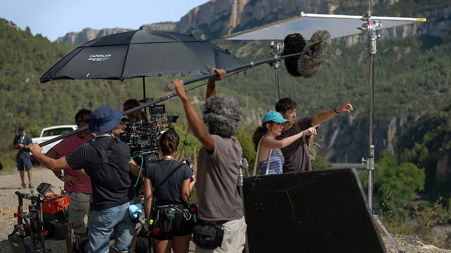 Filmteam beim Dreh im Freien vor bergiger Kulisse, mehrere Crewmitglieder stehen um Kamera und Tonangel, Regisseurin zeigt in eine Richtung, große Schirme spenden Schatten.