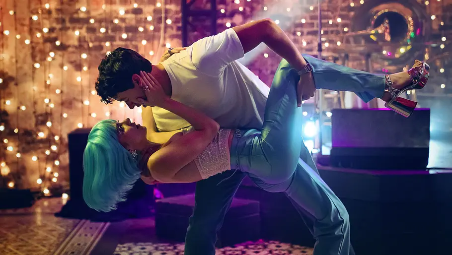 Ein Mann hält eine Frau mit türkisfarbener Perücke und glitzerndem Top in einer eleganten Tanzpose, während sie sich lachend ansehen. Sie tragen auffällige Kleidung und stehen auf einer Bühne mit funkelnder Lichterkette im Hintergrund, was eine festliche und lebendige Atmosphäre schafft.