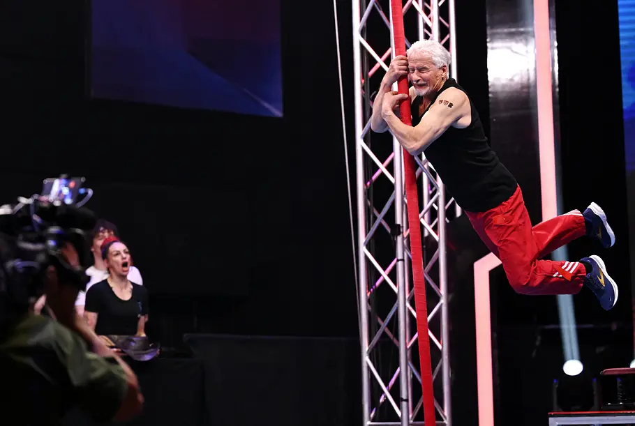 Peter Schlag: Ältester Teilnehmer bei Ninja Warrior Germany 2026 Peter Schlag hält sich mit angewinkelten Beinen an einer roten Stange fest. Er trägt ein schwarzes Oberteil und eine rote Hose.