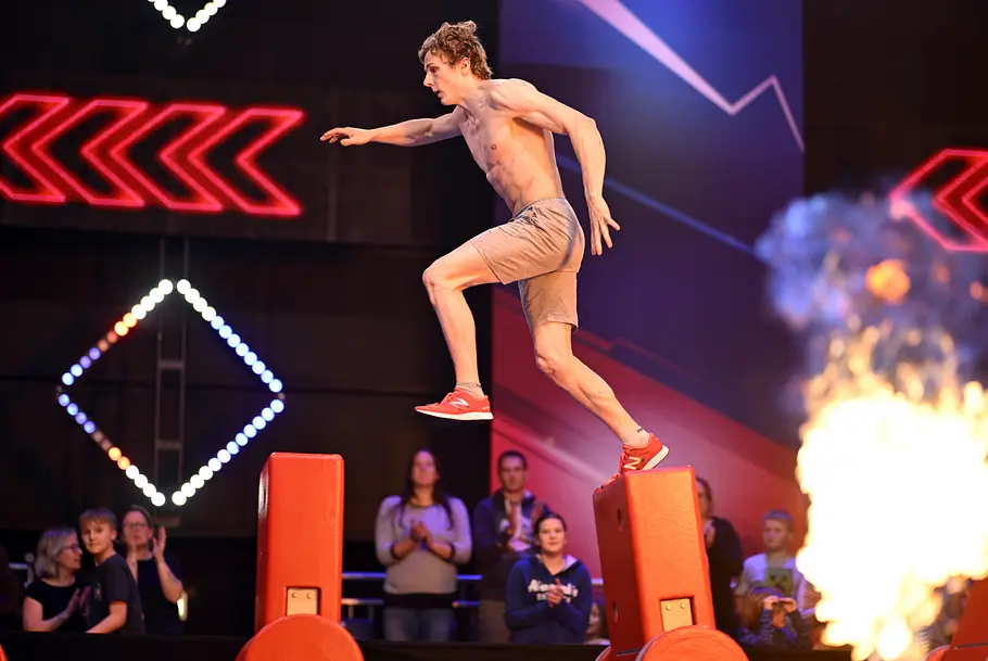 Philipp Göthert ist Teilnehmer bei Ninja Warrior Germany 2026 Philipp Göthert springt mit freiem Oberkörper zwischen roten Hindernissen. Rechts lodert eine Flamme, dahinter sitzt Publikum.