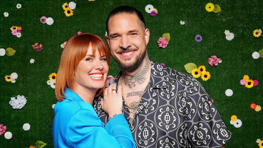 Pia Tillmann und Zico Banach Pia Tillmann im blauen Blazer und Zico Banach im gemusterten Hemd posieren lächelnd vor einer grünen Wand mit bunten Blumen.