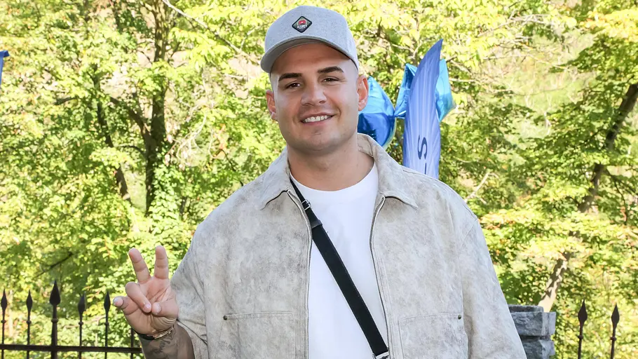 Pietro Lombardi trägt ein weißes Shirt, eine beige Jacke und eine graue Kappe. Er lächelt in die Kamera und zeigt mit der rechten Hand das „Peace-Zeichen.