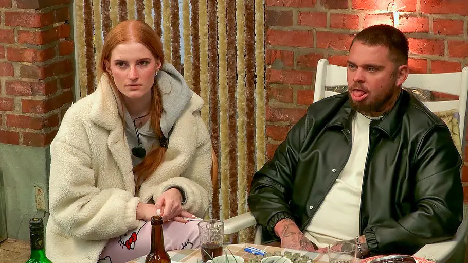 Laura und Nico Laura und Nico sitzen an einem Tisch in einem rustikalen Raum mit Backsteinwand; auf dem Tisch stehen Flaschen und Gläser, beide blicken ernst nach vorn.
